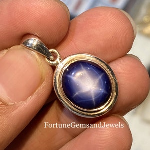May include: A silver pendant with an oval, deep blue gemstone. The gemstone has a star-like pattern in the center. The pendant is held in a person's hand. The text "FortuneGemsandJewels" is visible at the bottom.
