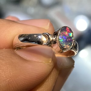 May include: A silver ring with an oval-shaped opal gemstone. The opal displays a vibrant array of colors, including red, green, and blue. The ring has a polished finish and a unique design on the band.