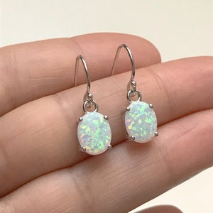 May include: A pair of oval-shaped opal earrings with a silver hook. The opals have a white base with iridescent flashes of green and blue. The earrings are set in a silver metal setting.