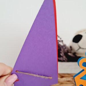 May include: Purple triangle banner with a red edge and a brown string attached. The banner is hanging from a string.