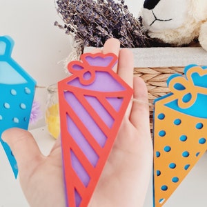May include: A colorful paper banner with a purple and red striped triangle, a blue triangle with white polka dots, and an orange triangle with blue polka dots. The banner is strung together with twine.