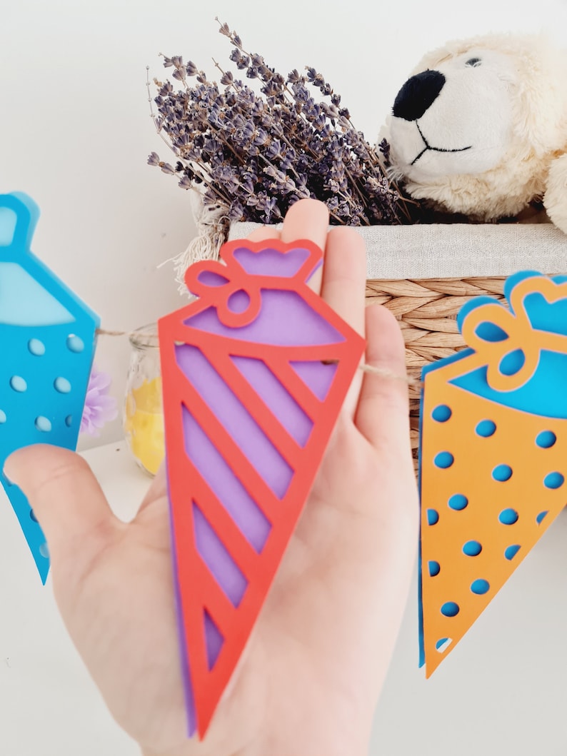 May include: A colorful paper banner with a purple and red striped triangle, a blue triangle with white polka dots, and an orange triangle with blue polka dots. The banner is strung together with twine.
