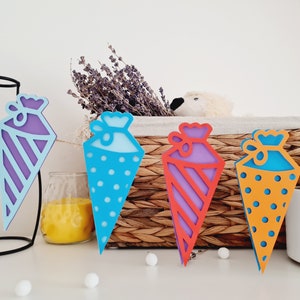 May include: A colorful paper garland with cone-shaped decorations. Each cone has a different pattern: purple and white stripes, blue with white polka dots, purple and red stripes, orange with white polka dots, and yellow with white stripes.