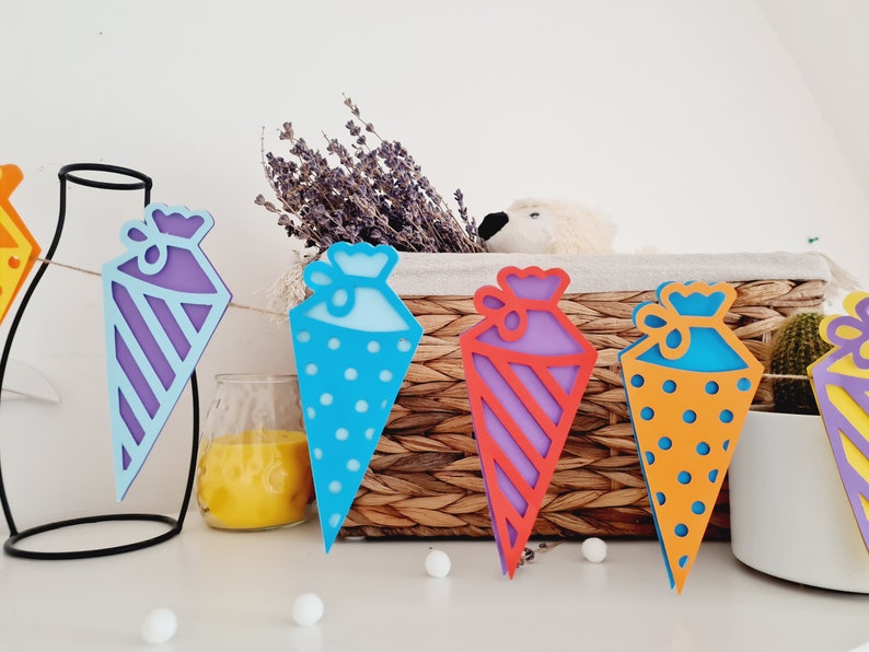 May include: A colorful paper garland with cone-shaped decorations. Each cone has a different pattern: purple and white stripes, blue with white polka dots, purple and red stripes, orange with white polka dots, and yellow with white stripes.