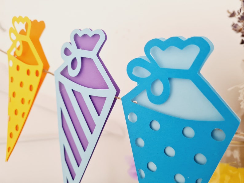 May include: A colorful paper garland with three pennants shaped like cones. The pennants are decorated with polka dots, stripes, and a bow. The garland is perfect for a birthday party or other celebration.