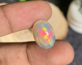 Può includere: Una gemma di opale di forma ovale con una base bianca lattea traslucida. La pietra mostra vibranti lampi di rosso, giallo e verde, creando un effetto di gioco di colori. La gemma è tenuta tra due dita.
