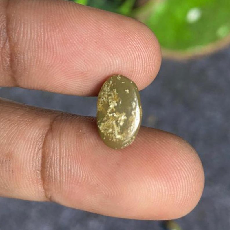 May include: An oval-shaped gemstone with a light olive-green hue and a pattern of lighter, textured inclusions. The polished surface reflects light, highlighting the stone's natural variations. This gemstone is suitable for jewelry making.
