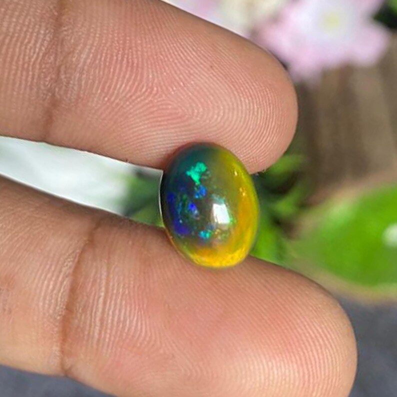May include: A vibrant, oval-shaped opal gemstone. The stone displays a mix of colors, including green, blue, and yellow, with a polished, reflective surface. The opal is held between fingers, showcasing its natural beauty and unique color play.
