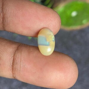 May include: An oval-shaped opal gemstone with a cream-colored base, a central band of pale blue, and yellow and green accents. The gemstone is held between two fingers, showcasing its polished surface and natural color variations.