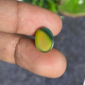 May include: An oval-shaped gemstone with a vibrant color gradient. The stone transitions from a bright yellow to a deep green hue. The polished surface reflects light, showcasing the stone's clarity and color variations. The gemstone is held between fingers.