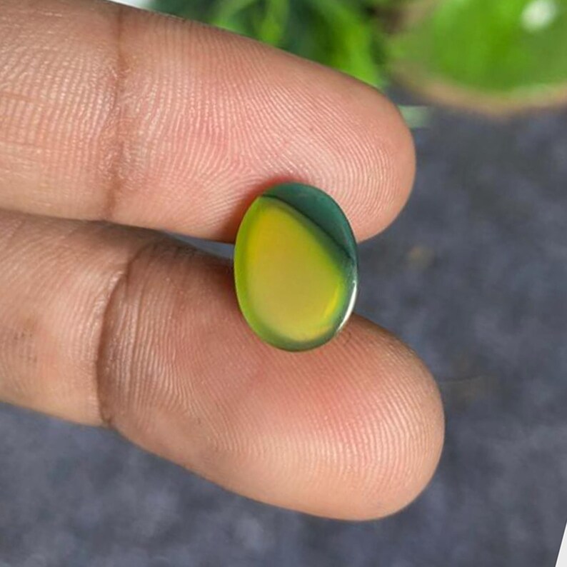 May include: An oval-shaped gemstone with a vibrant color gradient. The stone transitions from a bright yellow to a deep green hue. The polished surface reflects light, showcasing the stone's clarity and color variations. The gemstone is held between fingers.