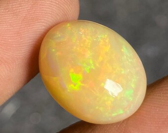 Può includere: Una gemma di opale lucidata, di forma ovale. La pietra ha un colore di base giallo pallido e cremoso con lampi iridescenti verdi e gialli. L'opale è tenuto tra le dita, mostrando la sua superficie liscia e riflettente.