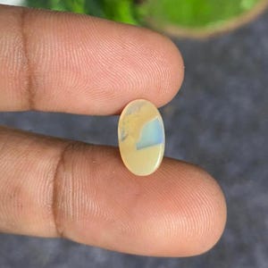 May include: An oval-shaped gemstone with a creamy yellow base color and a patch of pale blue and white. The gemstone is held between two fingers, showcasing its polished surface and unique color pattern. The gem is likely a cabochon cut.