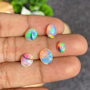 Puede incluir: Cinco piedras preciosas de ópalo coloridas con destellos iridiscentes de verde, azul, naranja y rosa. Los ópalos son de forma ovalada y varían en tamaño, mostrando una gama de patrones y colores naturales. Estas piedras son ideales para joyería.