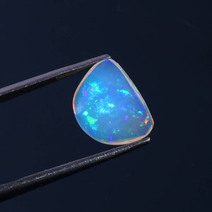 Puede incluir: Una piedra preciosa de ópalo azul claro con destellos iridiscentes de verde, rojo y morado. El ópalo se sostiene con unas pinzas sobre un fondo azul oscuro.