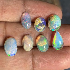 Puede incluir: Una colección de siete ópalos de varias formas, incluyendo ovalados y en forma de lágrima. Los ópalos muestran una gama de colores, incluyendo azul, verde, amarillo y rosa, con patrones iridiscentes. Las piedras preciosas se sostienen en una mano.
