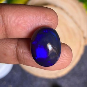 Puede incluir: Una piedra preciosa de ópalo negro pulido, de forma ovalada, con destellos azules y púrpuras vibrantes. La piedra se sostiene entre los dedos, mostrando su superficie reflectante y juego de colores. El ópalo es una piedra natural.