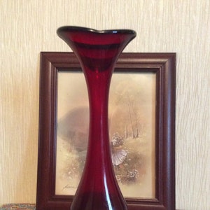 Peut inclure: Un vase en verre rouge rubis, haut et fin, avec une ouverture évasée. Le vase a une surface lisse et polie et une base en verre clair.