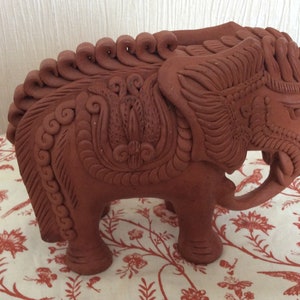 May include: A terracotta-coloured clay sculpture of an elephant featuring intricate, raised relief patterns along its back, head, and ears. The surface displays detailed swirling and floral-inspired decorative motifs carved into the clay body.