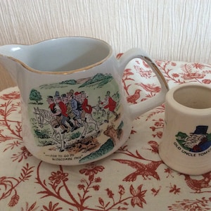 May include: A white ceramic pitcher and a small matching mug featuring colorful illustrations of characters from the song Widecombe Fair. The pitcher shows multiple people riding a horse with the text For to go to Widecombe Fair, while the mug depicts a man with the text Old Uncle Tom Cobley.