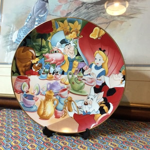 Può includere: Piatto decorativo con bordo dorato, raffigurante una scena di Alice nel Paese delle Meraviglie. Il piatto presenta Alice, il Cappellaio Matto e il Bianconiglio a una festa del tè, con un design colorato.