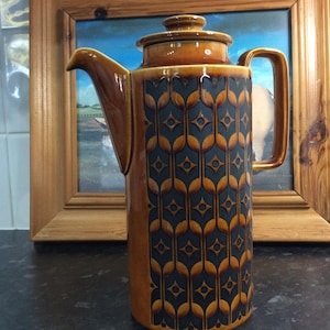 Peut inclure: Une cafetière en céramique marron avec un couvercle et une poignée. Le pot présente un motif répété de formes en forme de feuilles bleu foncé et dorées. Le pot est présenté devant une peinture encadrée.