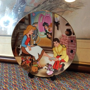 Può includere: Piatto decorativo raffigurante una scena di Cenerentola, con il Principe che prova la scarpetta di cristallo. Il piatto ha un bordo dorato ed è ambientato sullo sfondo di un camino in mattoni e una finestra con spettatori.