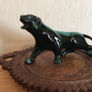 Può includere: Statuetta di pantera in ceramica verde scuro lucida con la bocca aperta, posta su un vassoio di legno intagliato. La pantera ha un design elegante e stilizzato, con un tocco di verde acqua sulle zampe. Il vassoio ha un bordo decorativo intagliato.