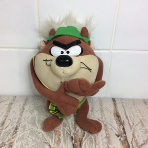 Può includere: Un peluche del Diavolo della Tasmania, un personaggio dei cartoni animati con pelliccia marrone, un cappello verde e capelli bianchi. Il giocattolo ha un'espressione arrabbiata e tiene le braccia incrociate. È un oggetto da collezione.