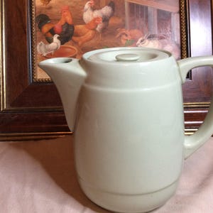 Op de afbeelding: Een lichtgroene keramische theepot met een deksel en een handvat. De theepot heeft een afgeronde body en een tuit. De achtergrond bevat een ingelijste schilderij van kippen en konijnen.