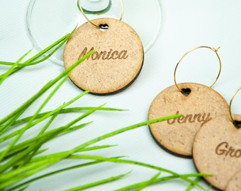 May include: Wooden wine glass charms with names engraved. The charms are round with a heart-shaped cutout and gold hoops. The name "Monica" is visible on one charm, with other charms in the background. Green grass-like decoration is in the foreground.
