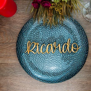 May include: A teal blue round plate with a textured surface. The name "Ricardo" is spelled out in gold letters on the plate.