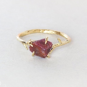 Könnte beinhalten: Ein goldener Ring mit einer strukturierten Ringschiene in Form eines dünnen Baumzweigs mit kleinen Ästchen. Der Ring ist mit einem einzelnen, rauen, dunkelroten Edelstein besetzt, der von vier Krappen gehalten wird. Der Text ADALGO handmade jewelry ist sichtbar.