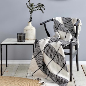 Könnte beinhalten: Eine schwarz-weiße karierte Wolldecke mit Fransen an den Rändern, die über einen schwarzen Holzstuhl gelegt ist. Die Decke ist aus einem weichen, strukturierten Material gefertigt und hat eine dezente Waffelstruktur.