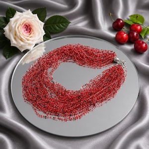 May include: A multi-strand necklace composed of numerous tiny red and black beads. The necklace features a silver-toned metal clasp and is arranged in a circular shape on a reflective surface, showcasing a vibrant array of small, uniform beads.