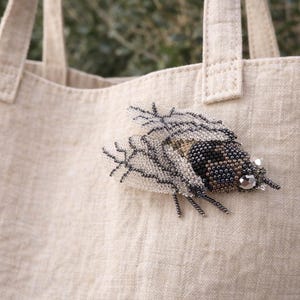 Könnte beinhalten: Eine bestickte Insektenbrosche in Schwarz-, Grau- und Beigetönen ist an einer beigen Tragetasche befestigt. Die Brosche hat ein detailliertes Design mit einem Kristallakzent. Die Tragetasche hat zwei Griffe und ist aus einem strukturierten Stoff gefertigt.