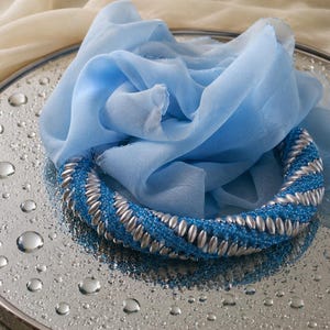 May include: A circular necklace featuring a twisted design of bright blue seed beads and metallic silver oval beads. The necklace is paired with a light blue sheer fabric scarf, creating a layered appearance with contrasting textures and colors.
