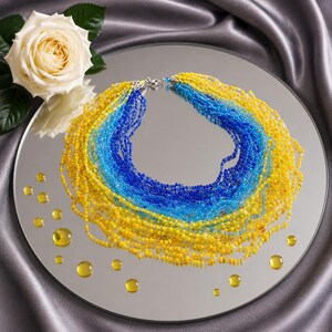 May include: A multi-strand beaded necklace featuring a vibrant gradient of yellow, light blue, and dark blue glass beads. The necklace is arranged in a circular shape with a silver-toned metal clasp closure at the top centre of the strand arrangement.