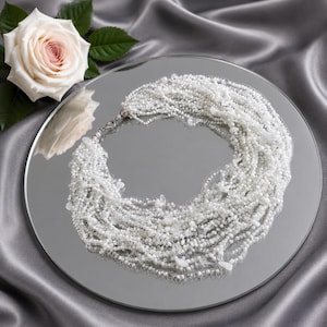 May include: A multi-strand necklace composed of numerous small white beads and irregular clear crystal-like accents. The necklace features a silver-toned metal clasp and is arranged in a circular shape on a reflective surface next to a white rose.