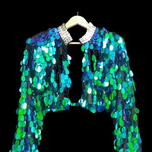 Puede incluir: Una chaqueta corta de lentejuelas verde azulado y verde con un cuello de pedrería.