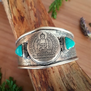 May include: Silver bracelet featuring a turquoise stone and a circular design depicting a seated Buddha. The bracelet is inscribed with Tibetan script.
