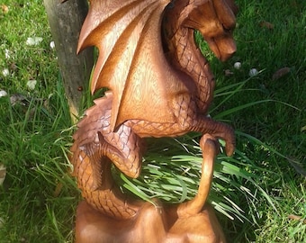 May include: A hand-carved wooden dragon sculpture with detailed wings and scales. The dragon is perched on a rock-like base and is painted in a warm brown color.