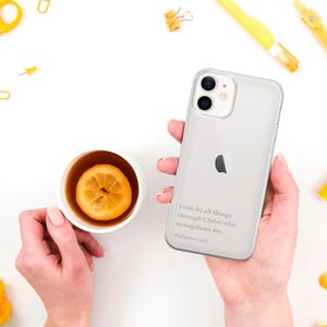 以下が含まれることがあります： Appleのロゴと「私は私を強くするキリストを通してすべてのことができます。ピリピ人への手紙4:13」というテキストが入った透明なiPhoneケース。もう一方の手には、レモンスライスが入ったお茶のカップが持たれています。
