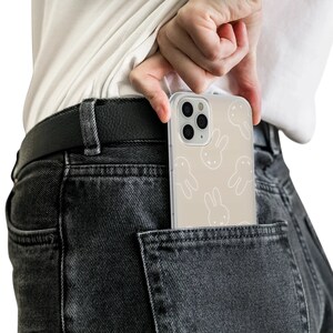 May include: A clear phone case with a light brown background and white bunny rabbit silhouettes. The case is in the back pocket of a pair of dark gray jeans.