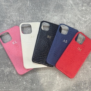 May include: Five phone cases with a textured crocodile skin pattern in pink, white, black, navy blue, and red. Each case features a square camera cutout and gold-coloured embossed initials: KL, AF, ES, AS, and JM, respectively, on the back surface.