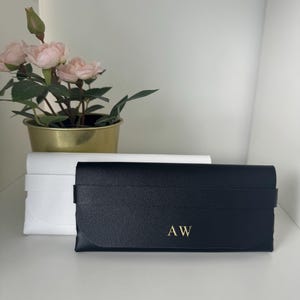Könnte beinhalten: Ein schwarzes, rechteckiges Brillenetui mit strukturierter Oberfläche und den goldenen, geprägten Initialen AW auf der Vorderseite, platziert vor einem weißen Brillenetui im gleichen Design und einem goldenen Topf mit künstlichen rosa Rosen.