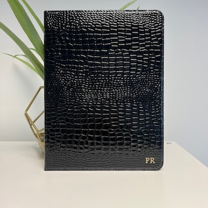 Puede incluir: Cuaderno de cuero negro con un patrón de cocodrilo en relieve y las iniciales doradas "PR" en la esquina inferior derecha.