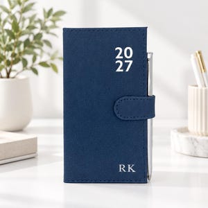 May include: A navy blue pocket planner with a magnetic closure strap and a silver pen inserted into the side loop. The front cover features the white printed text 20 27 at the top and the initials RK in white at the bottom right corner.