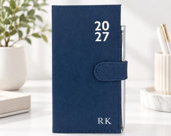 May include: A navy blue pocket planner with a magnetic closure strap and a silver pen inserted into the side loop. The front cover features the white printed text 20 27 at the top and the initials RK in white at the bottom right corner.