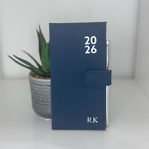May include: A navy blue leatherette journal with the year "2026" printed in white. The initials "RK" are printed in white at the bottom. The journal has a snap closure and is standing upright next to a small potted plant.